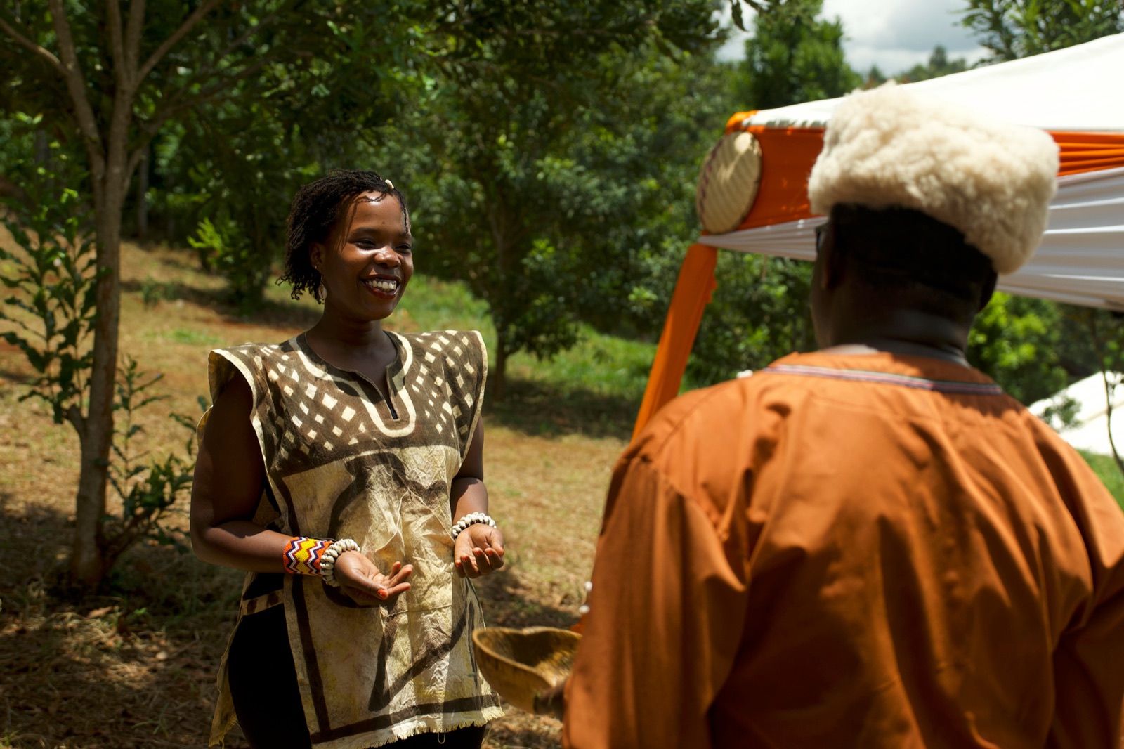 Wangechi Mwangi laughing in conversation with an elder in traditional dress, outdoors among trees.